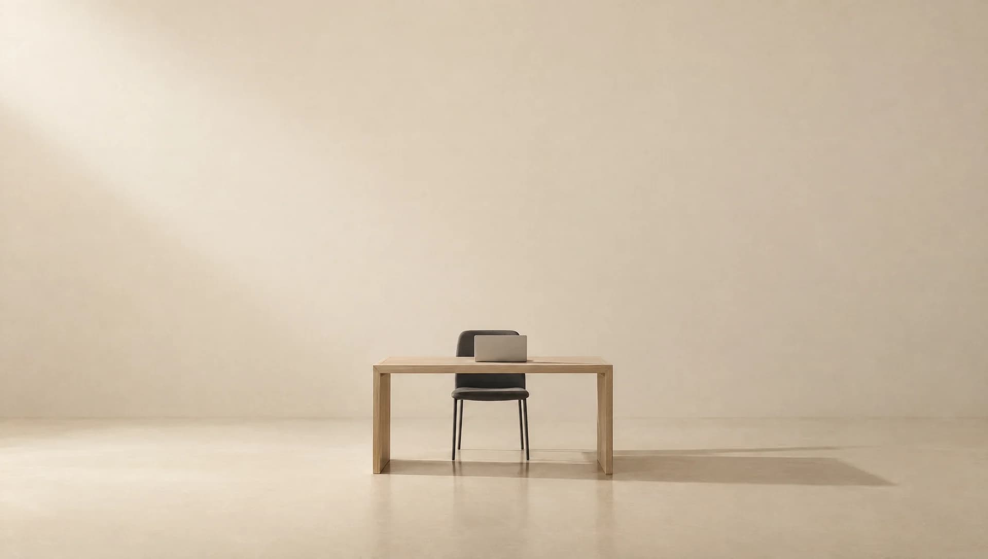 A single desk with a laptop and an empty chair, alone in a large warm cream room.